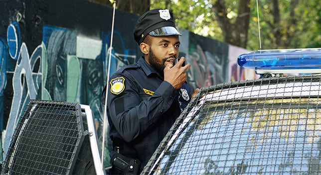 A uniformed police officer stands next to his vehicle and speaks into the radio