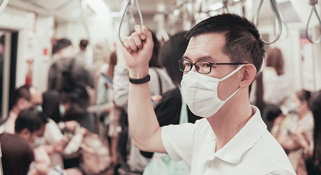 A man with glasses stands in a subway car wearing a white cloth surgical mask over his nose and mouth
