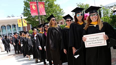 A line of University of Southern California students at their graduation ceremony with the person in front smiling and holding a sign that reads, “Geographic Information Science and Technology”