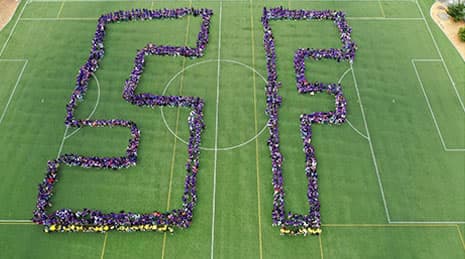 A top-down aerial view of a grass sports field with many people standing together to form the letters “S” and “F”