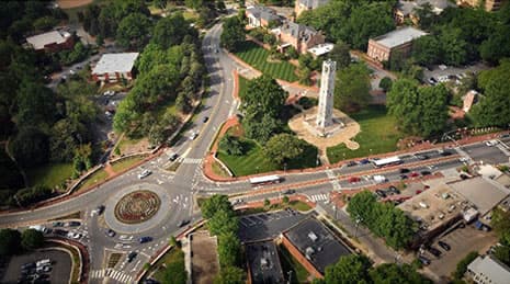 A top-down aerial view of city roads with a roundabout, a monument, buildings, and trees surrounding the area