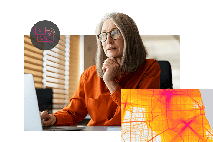 Eine Frau in einer orangefarbenen Bluse sitzt in einem hell beleuchteten Büro am Schreibtisch und arbeitet am Laptop.