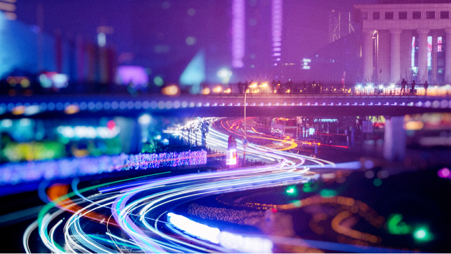 A long-exposure nighttime view of a city shows a pedestrian bridge over a busy highway with streaks of white and orange light