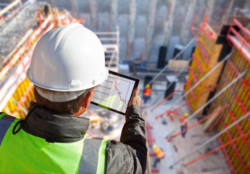 A construction worker wearing a hard hat and safety vest overlooks a construction site while working on a tablet.