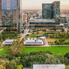 An aerial photo of a tree-filled green public city park surrounded by modern skyscrapers under a hazy orange sunset sky