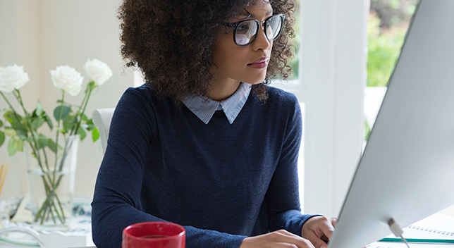 Mujer joven con gafas en un escritorio mirando el monitor grande plateado del PC