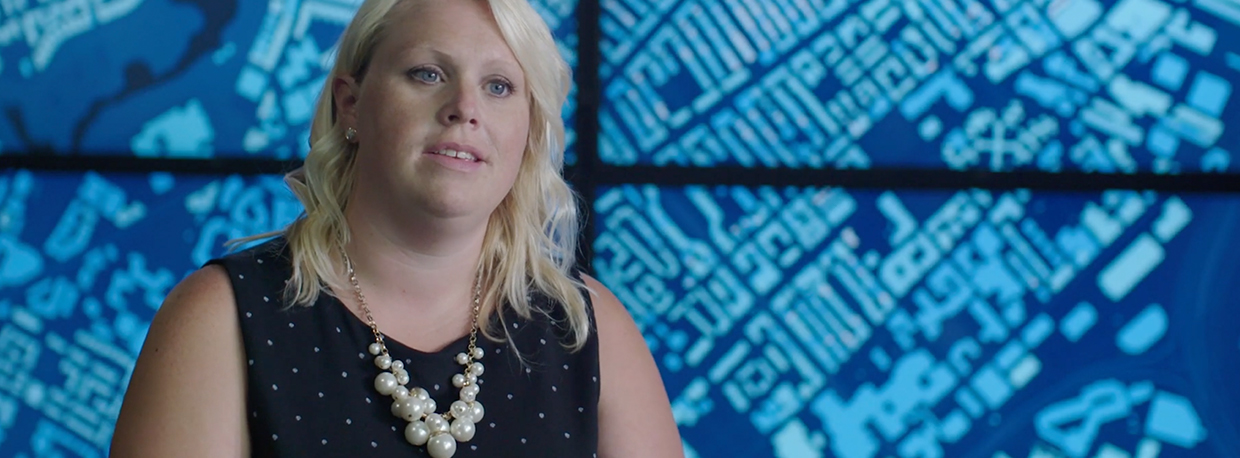 A woman wearing a black polka dot dress and a pearl necklace is seated in front of a blue digital map display.