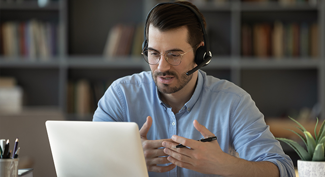 A man wearing a hands-free headset with a microphone talks while looking at a laptop screen.