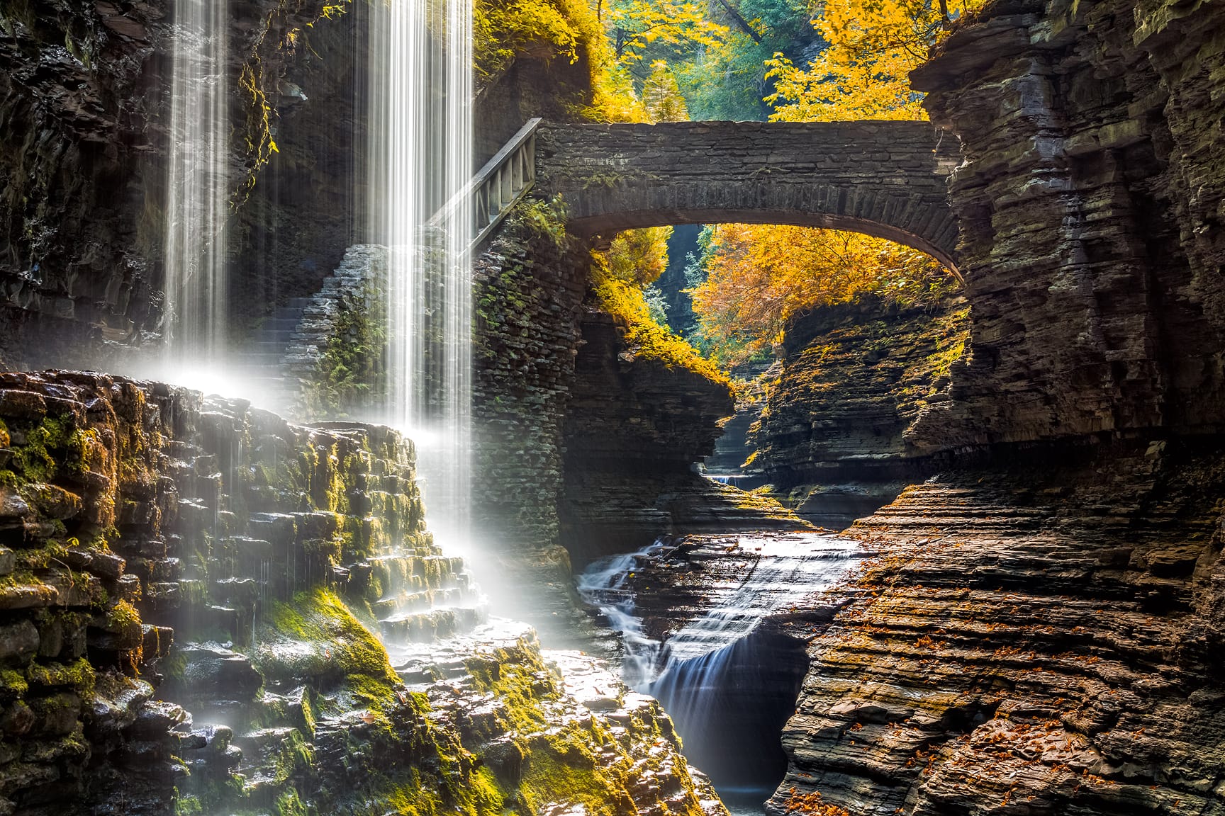 A picturesque stone bridge arches over a cascading waterfall in a rocky gorge. Sunlight filters through autumn foliage, illuminating the water and moss-covered rocks.