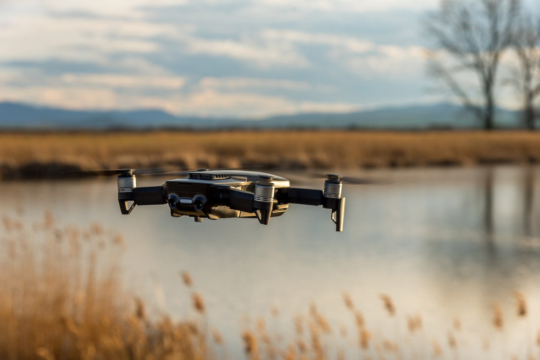 A small quadcopter drone hovers in mid-air above a calm lake, surrounded by tall grasses and distant mountains. 