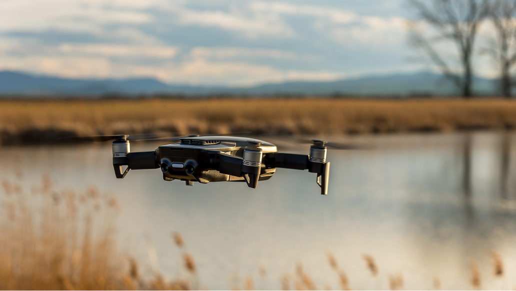 A small quadcopter drone hovers in mid-air above a calm lake, surrounded by tall grasses and distant mountains. 