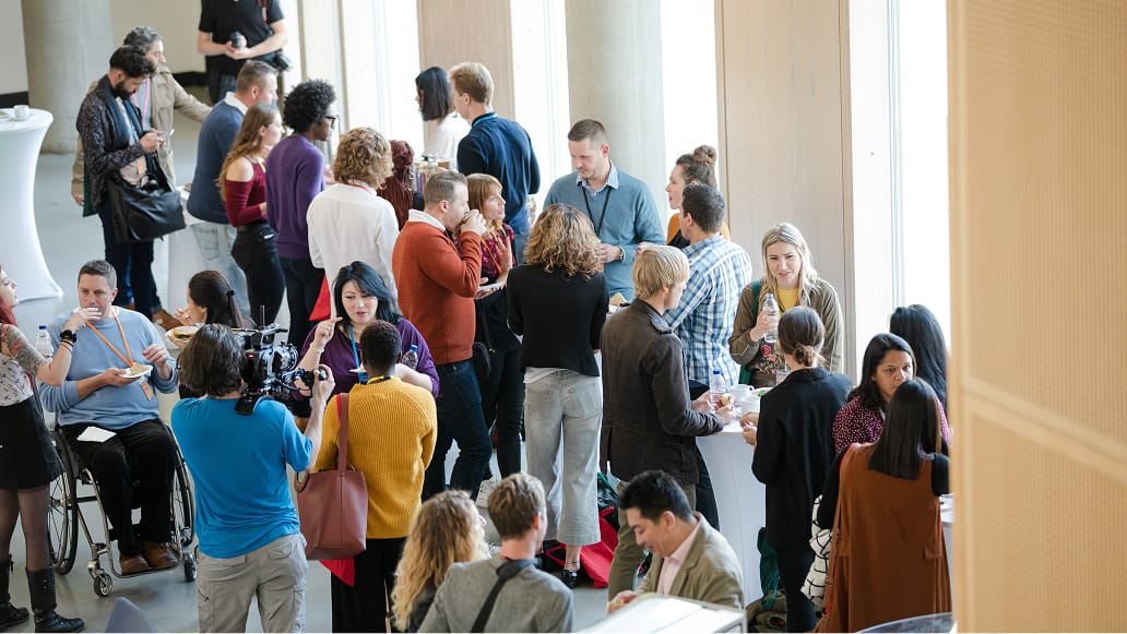 A large group of adults gathers in a bright, modern indoor space, engaging in conversation and networking. 
