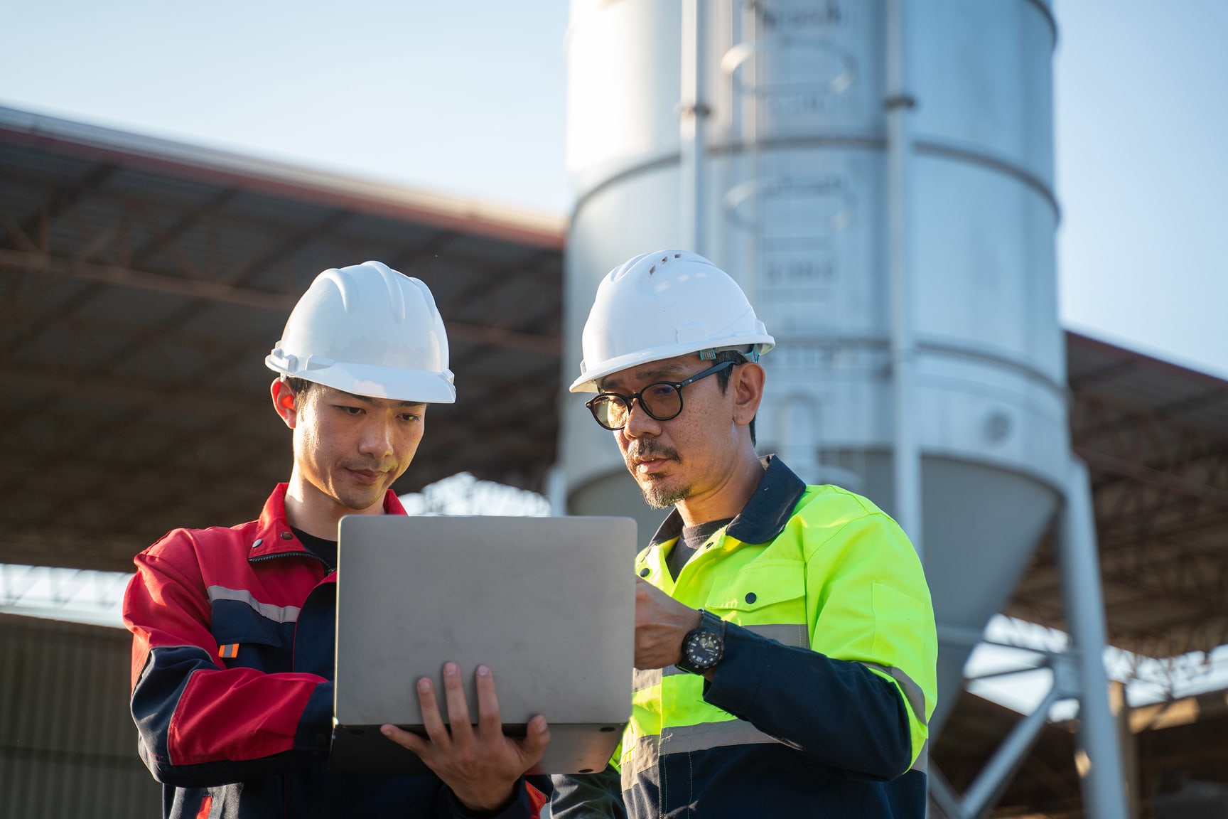 Two construction workers review a laptop at an outdoor industrial site.
