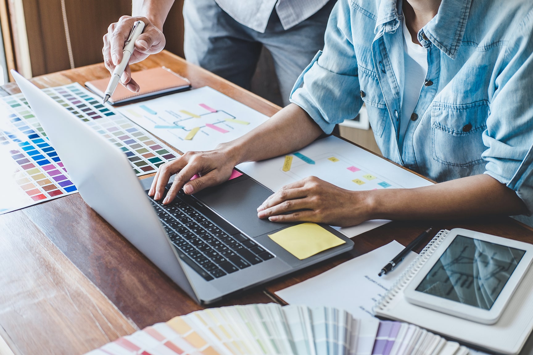 Two designers work together at a wooden desk, reviewing color swatches beside an open laptop