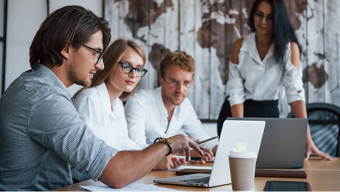 A group of professionals gathers around a table with laptops and documents, engaged in a collaborative discussion.