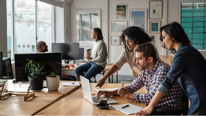 A group of colleagues gathers around a laptop in a bright, contemporary office environment. 