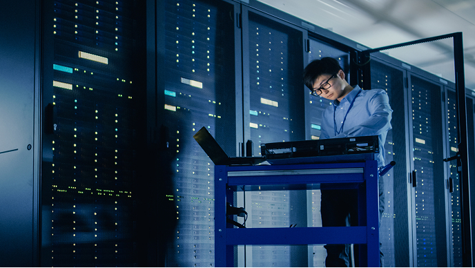 A person is seen operating a laptop on a mobile workstation in a data center filled with server racks.