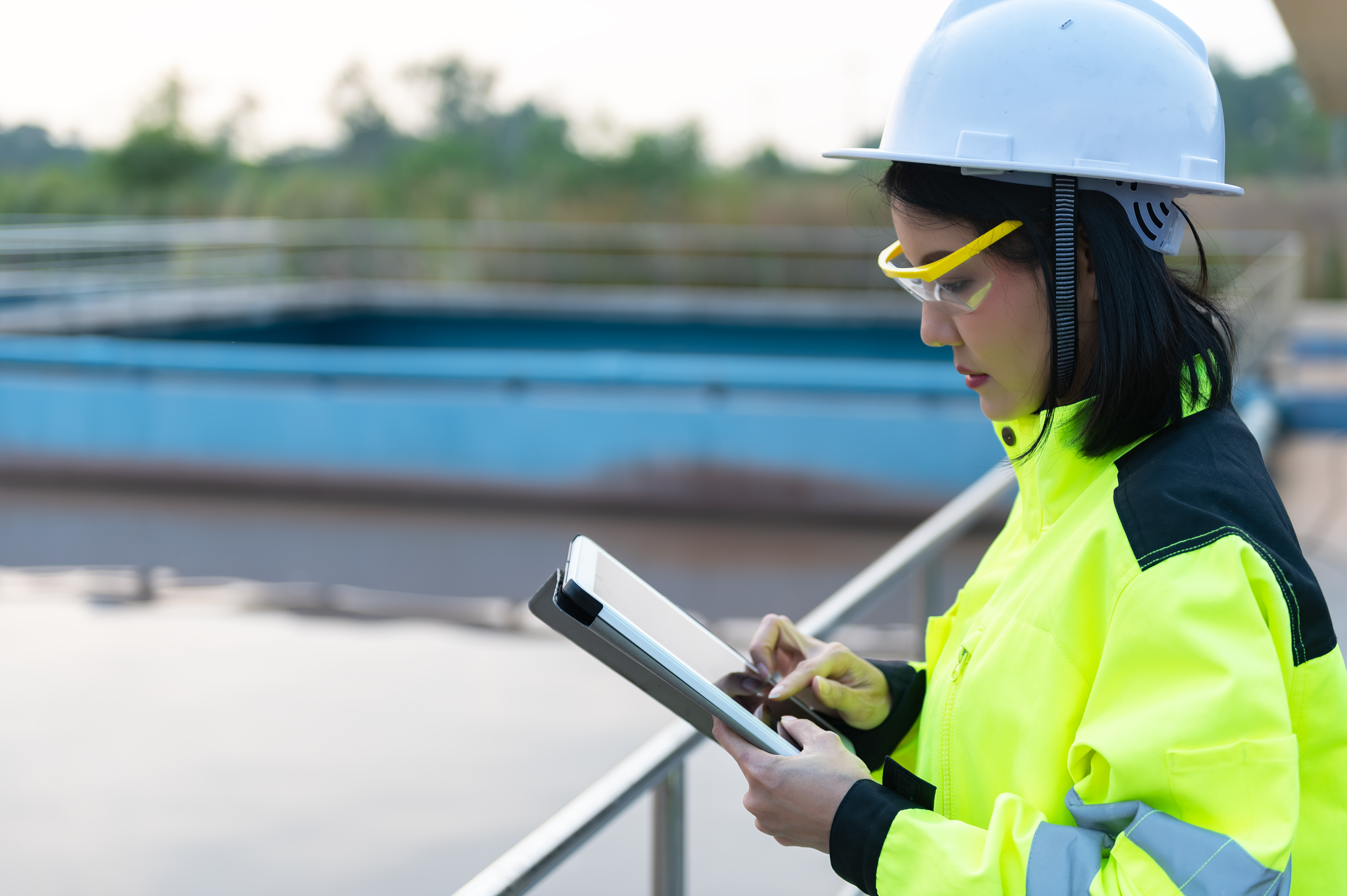 Female utility worker in  a white hardhat, safety glasses, and a yellow and black safety jacket is outdoors, holding a tablet 