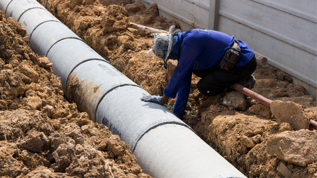 Male worker wearing a hard hat, long-sleeve shirt, and jeans kneels beside an open ditch, with a hand on a large water pipe. 