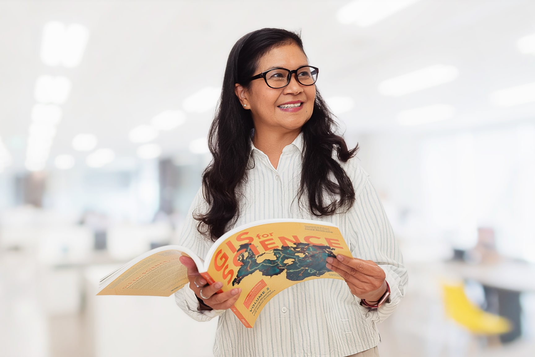 A woman stands in a bright, modern office environment holding and reading a magazine titled 'GIS for Science.' 