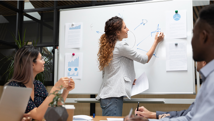 Woman standing at a whiteboard diagramming the parts of a system while another woman seated looks on with interest.