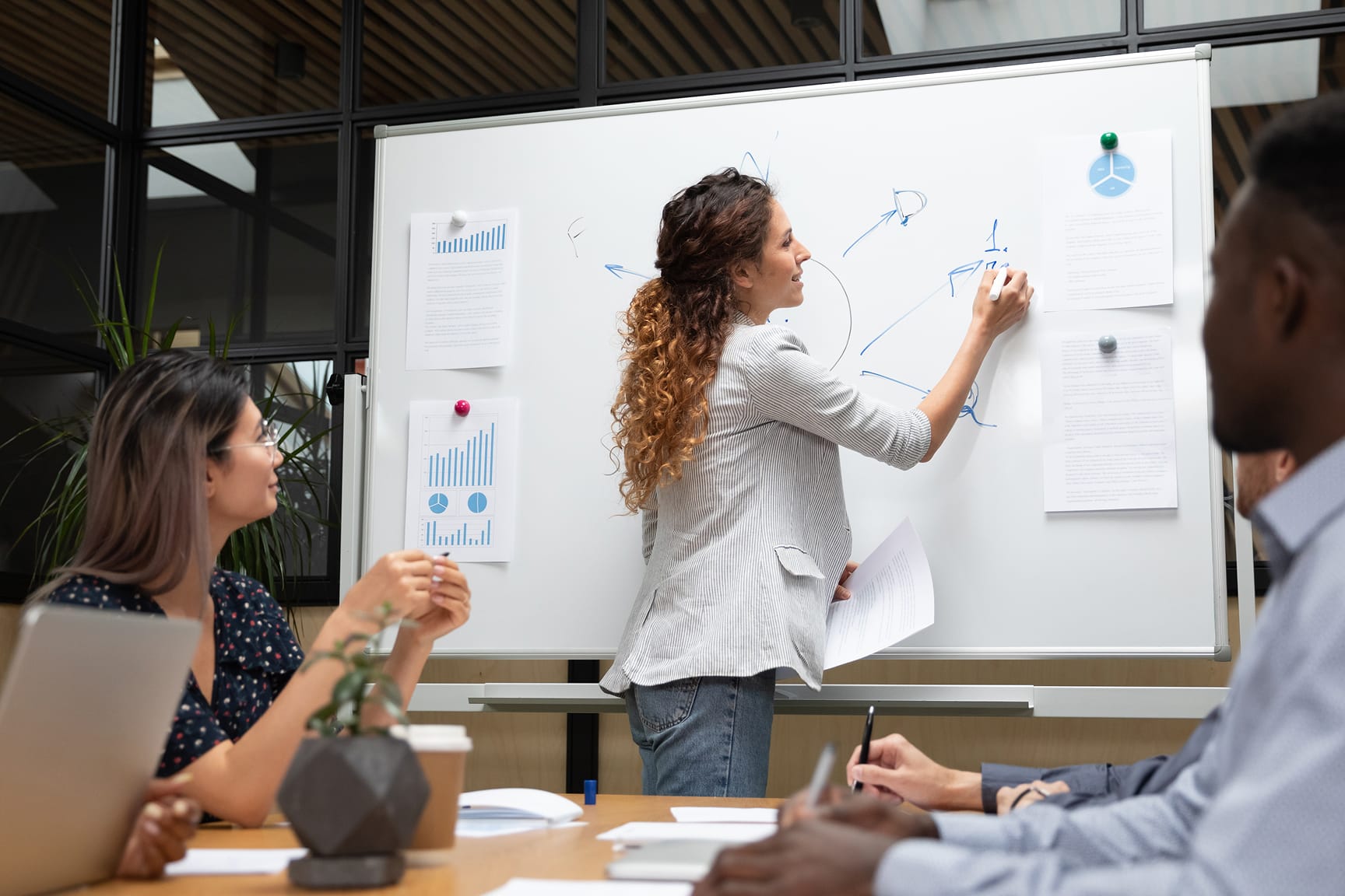 A collaborative team meeting in a modern office setting, featuring a woman presenting ideas on a whiteboard