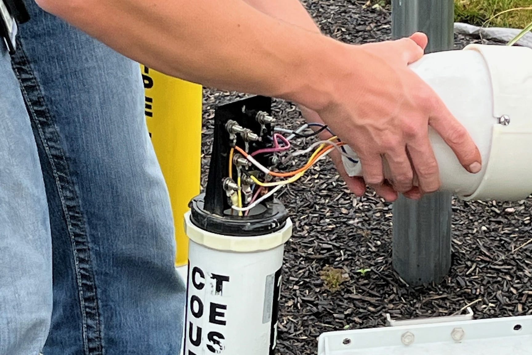A person is working on an open cylindrical electrical junction box outdoors, connecting or inspecting multiple colored wires.
