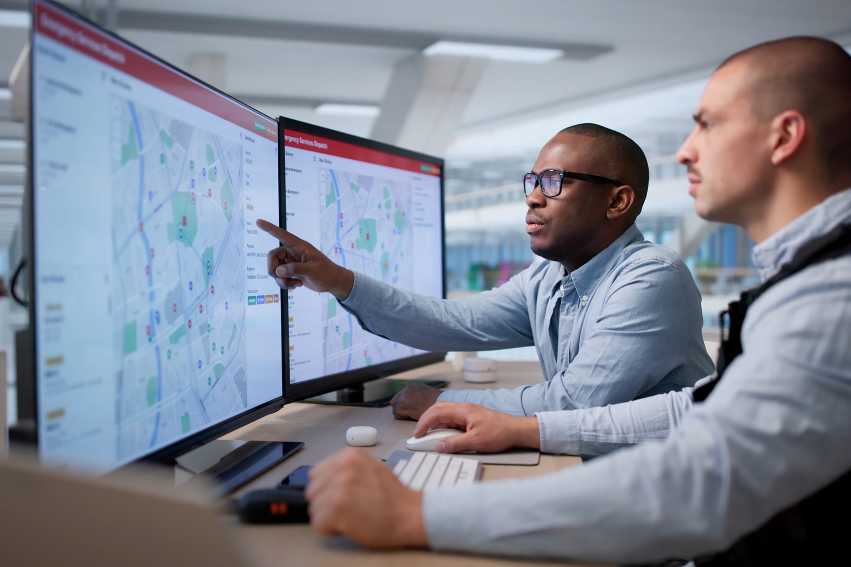 Two professionals sit at a desk analyzing detailed city maps on dual computer monitors