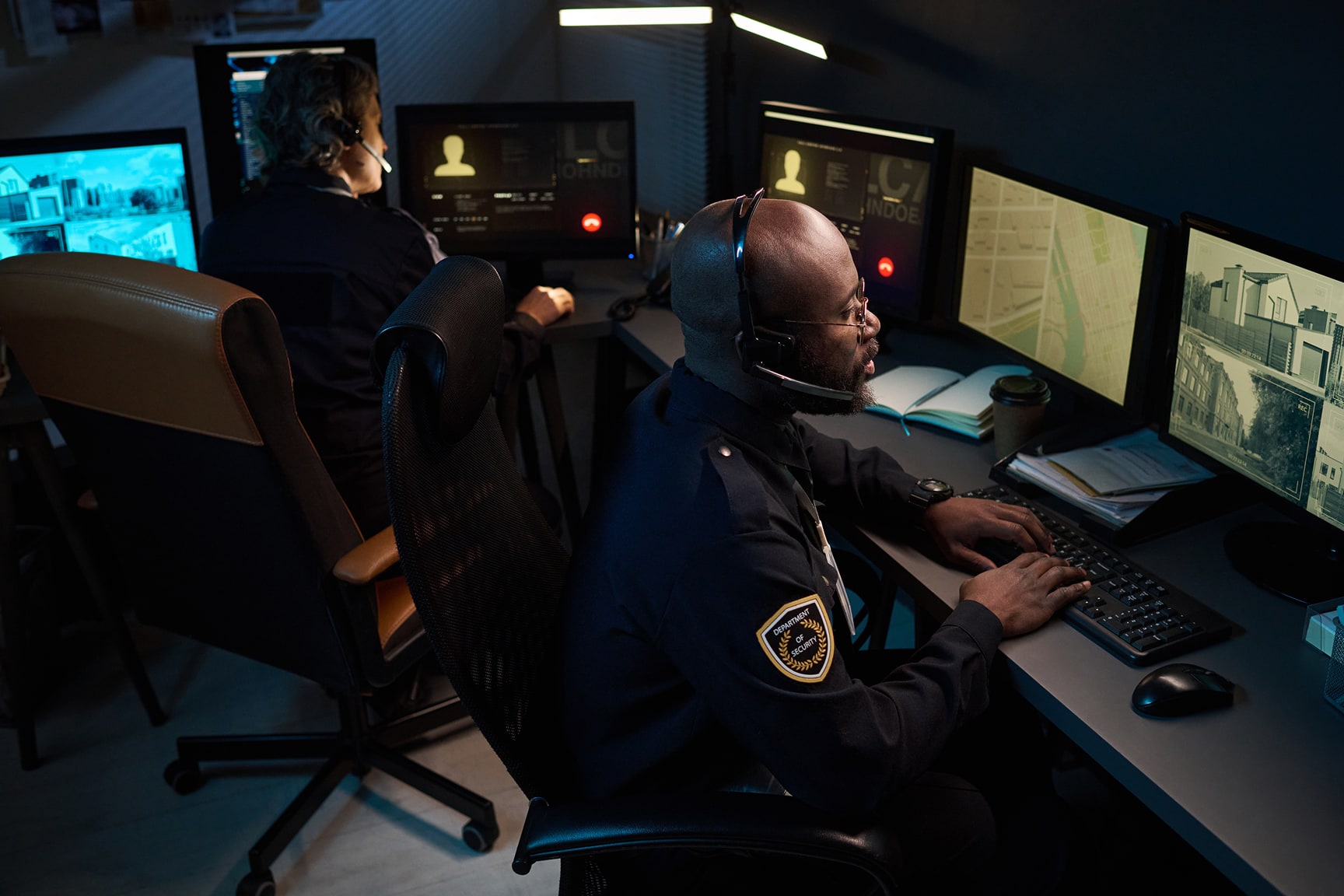 Two security officers sit at computer workstations in a dark control room, monitoring multiple surveillance screens