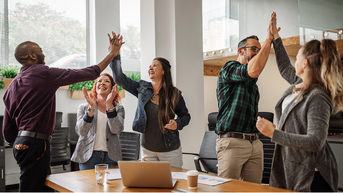 Image of several individuals standing up from a table with open laptops, engaging in a group high five with hands raised in the air.