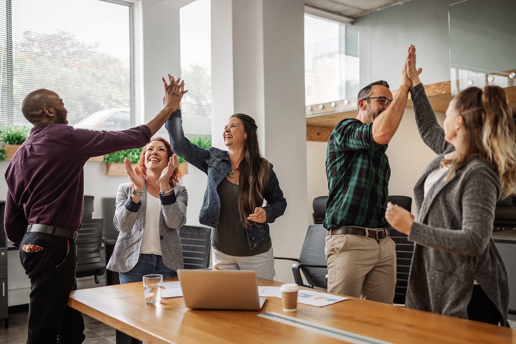 A group of diverse colleagues in a modern office setting enthusiastically giving high fives around a conference table. 