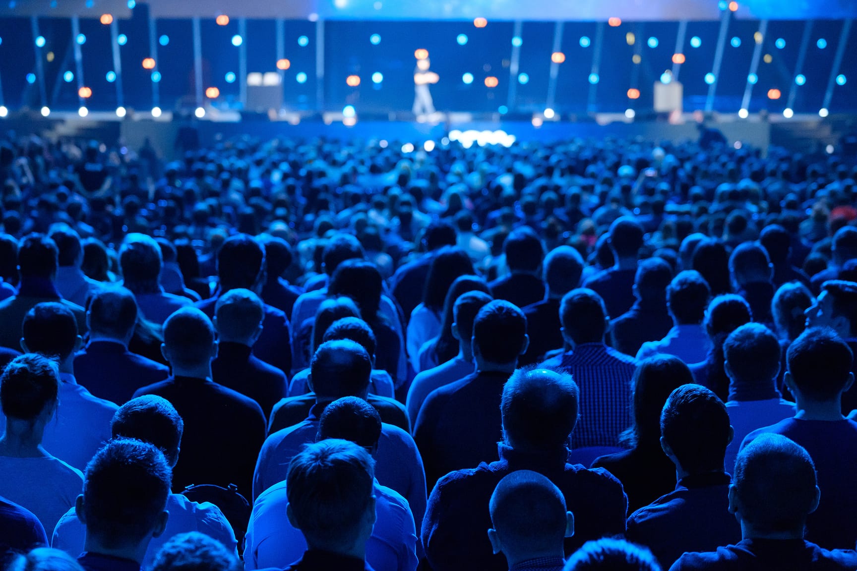 The image shows large audience of people looking at a stage in a dark setting.