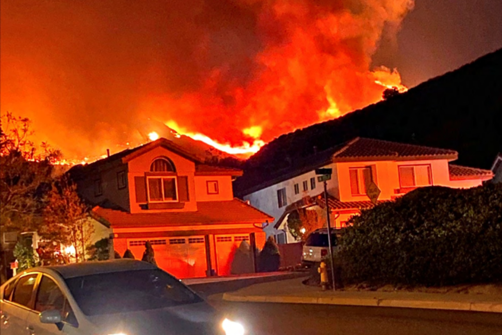A large wildfire rages on a hillside directly behind a row of suburban houses at night