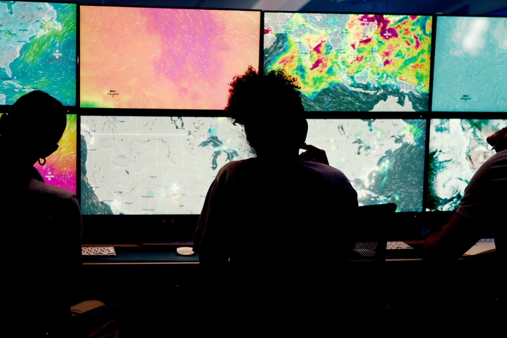 Three silhouetted figures keep watch over a bank of monitors featuring real-time maps. 