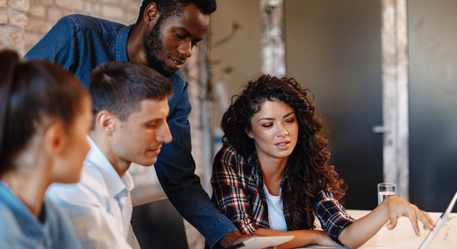 Four people working around a table and pointing to a computer