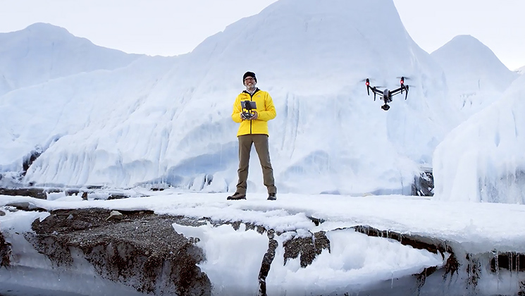 A man in a yellow jacket flies a drone at Mount Everest, two hikers wearing red and yellow jackets look out over a snowy canyon
