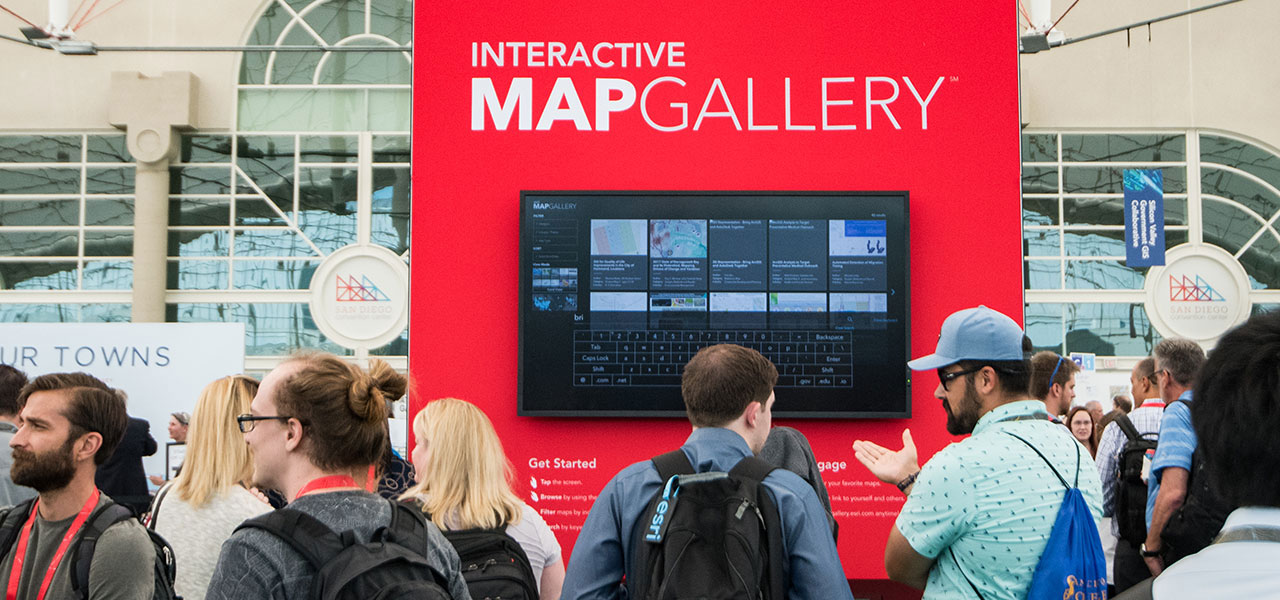 A crowd of people standing in front of a large red stand displaying Esri’s Interactive Map Gallery on a monitor
