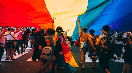 A group of people marching in a parade underneath a large rainbow flag 