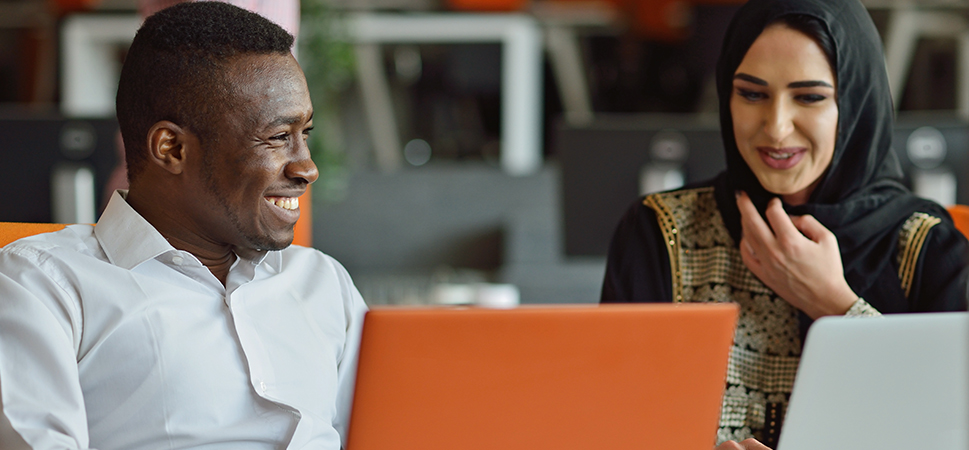 Two diverse people seated and laughing looking at a laptop screen