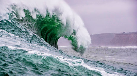 Photo of a green ocean wave curling in the foreground with a hilly shore in the background beneath a foggy sky