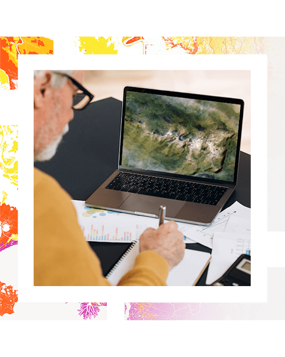 A photo of a geographer sitting at a table working on a laptop, which displays a satellite photo of a green mountain range