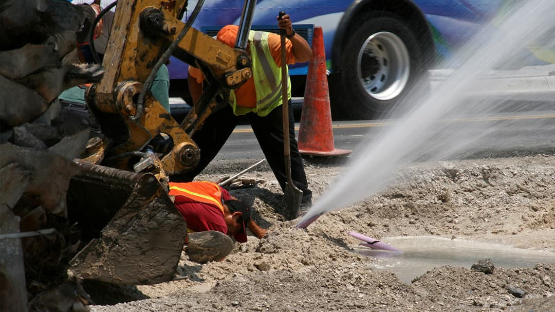 Two construction workers are seen repairing a water pipe leak on a busy street.
