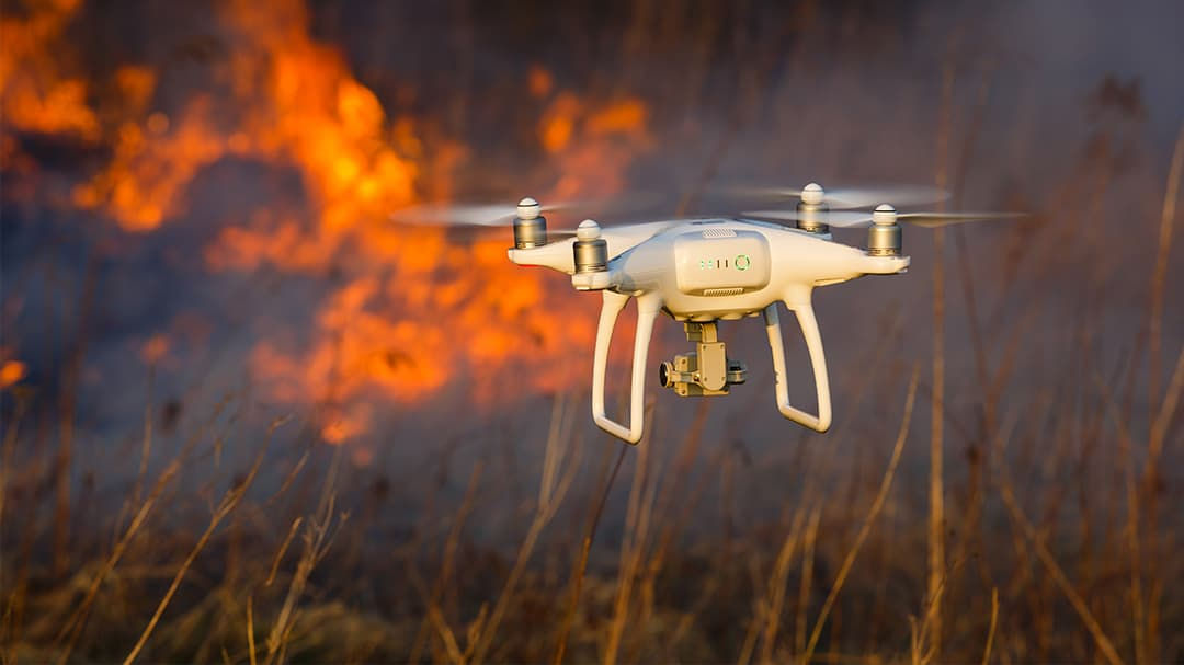A white quadcopter drone hovers in the foreground of a wildfire scene, surrounded by dry grass and intense flames. 