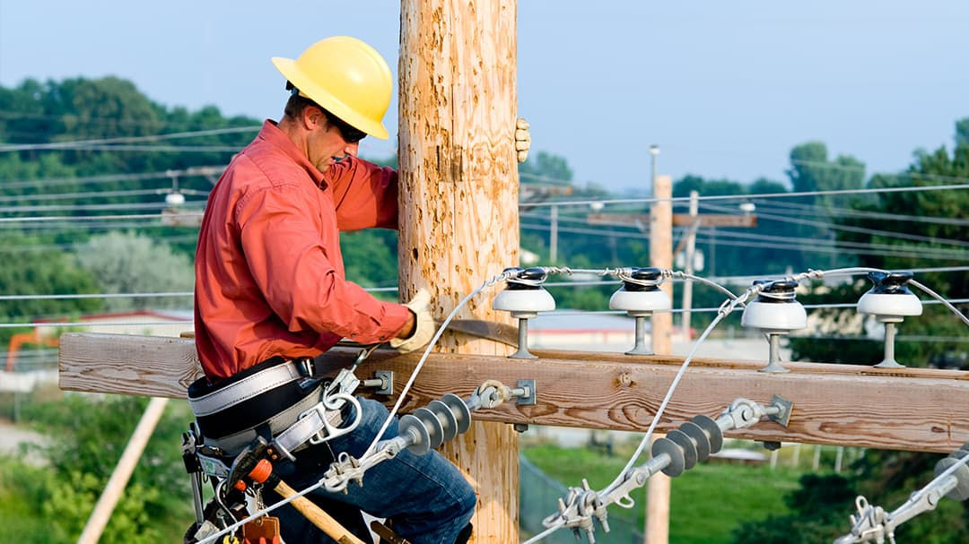 A utility worker wearing a yellow safety helmet and red shirt is repairing power lines on a wooden pole.