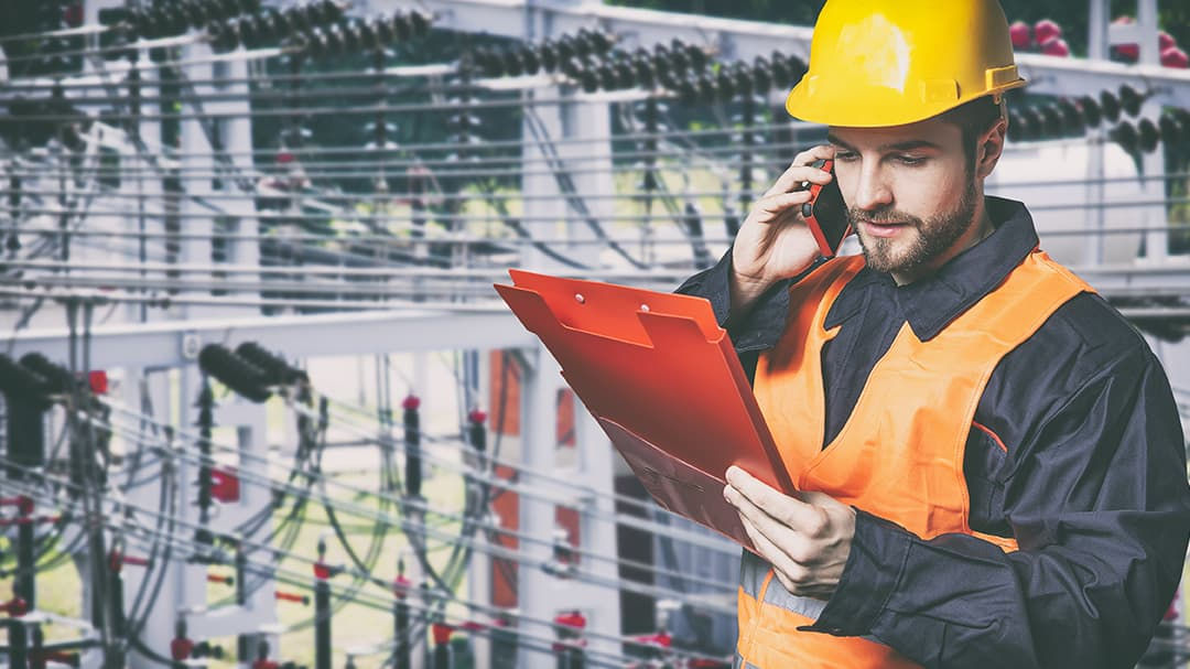 An engineer wearing a yellow hard hat and orange safety vest holds a red clipboard while inspecting electrical power lines.