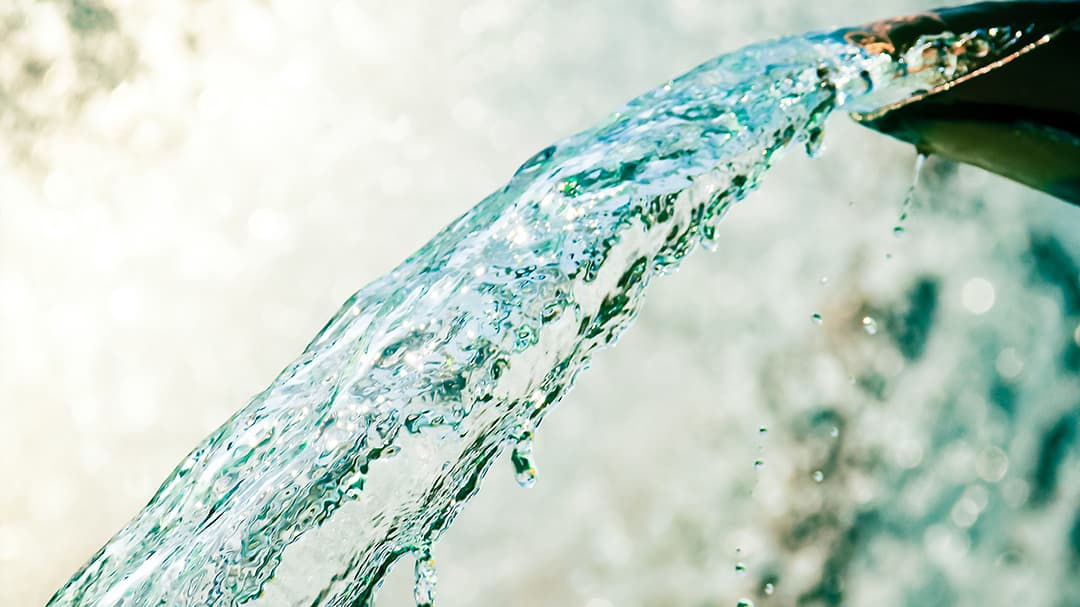 A close-up view of clear water flowing from a metal spout, captured in an outdoor setting.