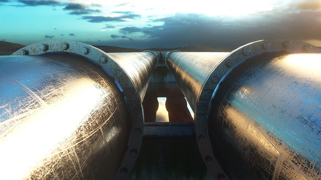 Two large metallic pipelines stretch across a rural setting under a dramatic sunset sky.