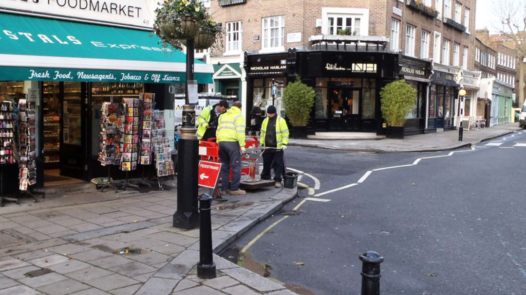 A group of workers in high-visibility jackets are seen on a city street corner near a food market and other shops. 