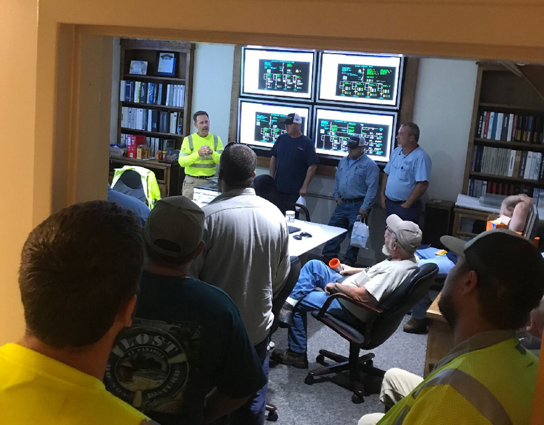 A group of workers gathers in a control room for a meeting.