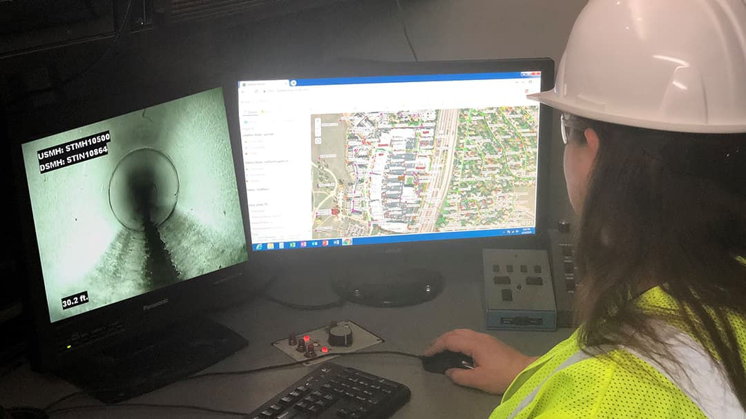 A person wearing a white hard hat and high-visibility vest works at a desk with dual monitors. 